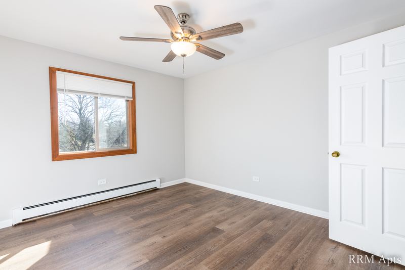 Bedroom with wood-look floor, ceiling fan, window with bare trees and a white door, Pioneer Court apartment 900-3D