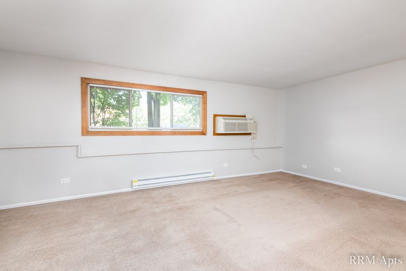 Living room with beige carpet, wide window with trees outside and a wall air conditioner, Pioneer Court apartment 918-1E