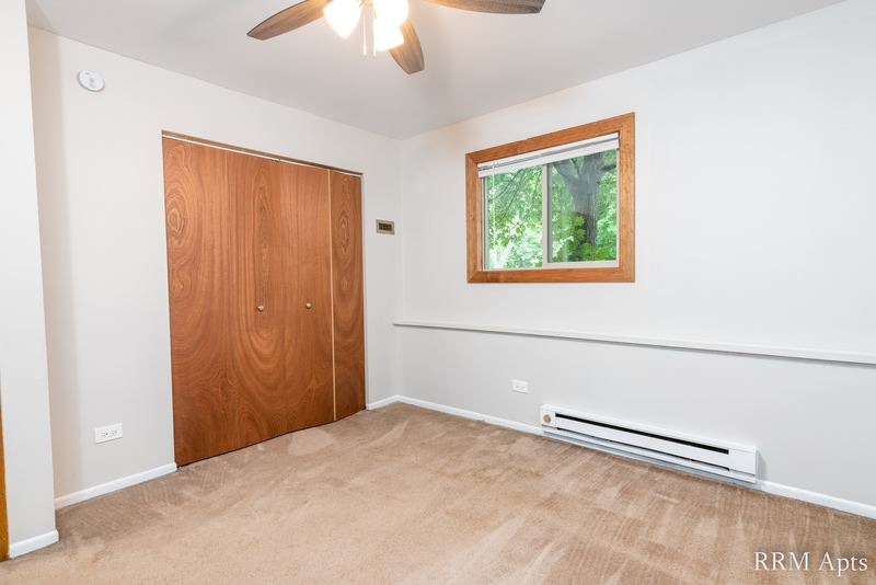 Bedroom with beige carpet, wood folding closet doors, window onto trees and a ceiling fan, Pioneer Court apartment 918-1E