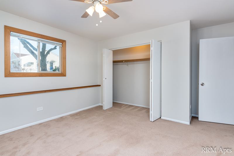 Bedroom with beige carpet, ceiling fan, window and open white folding closet doors, Pioneer Court apartment 918-1F