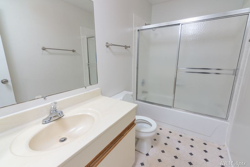 Bathroom with cream vanity, mirror, toilet and glass-door tub, patterned tile floor, Pioneer Court apartment 918-1F