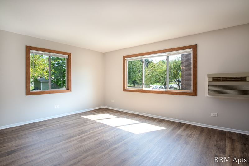 Living room with wood-look floor, two windows with green trees and a wall air conditioner, Pioneer Court apartment 918 2A
