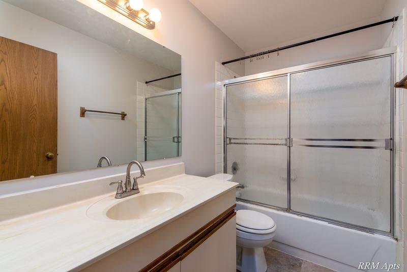 Bathroom with cream vanity, wide mirror, sliding glass tub doors, toilet and an oak door, Pioneer Court apartment 918 2E