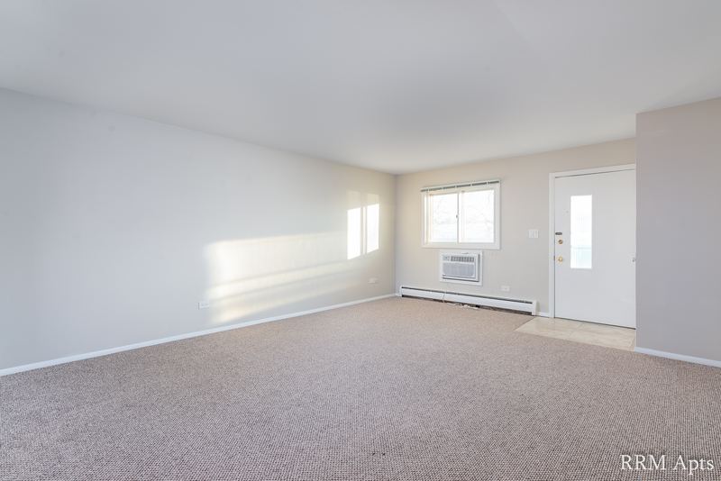 Living room with beige carpet, wall air conditioner, baseboard heater and entry door with window, Pistakee Road apartment 1A