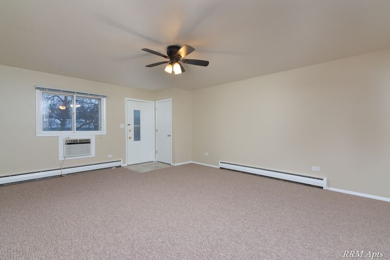 Living room with beige carpet, ceiling fan, wall air conditioner under a window and entry door, Pistakee Road apartment 1C