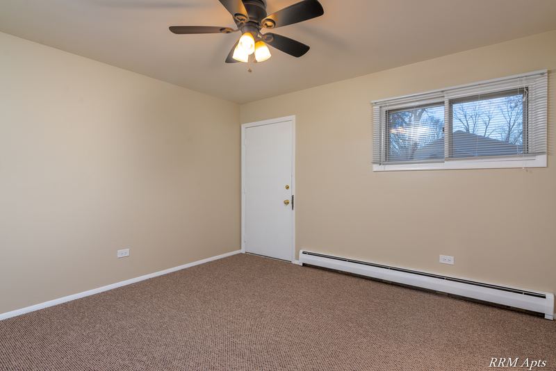 Empty room with beige carpet, ceiling fan, small window, white door and baseboard heater, Pistakee Road apartment 1C