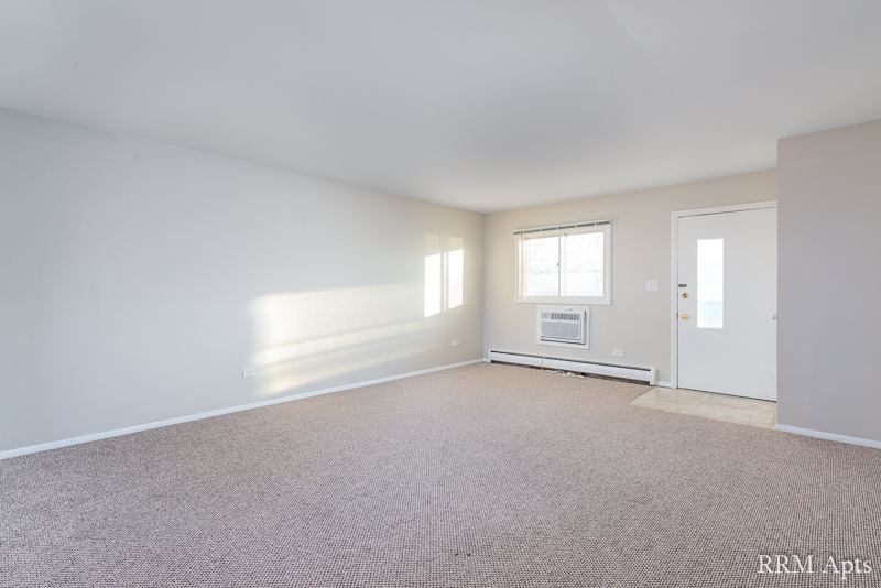 Carpeted living room with window, wall air conditioner and front door, Pistakee Road apartment 1D