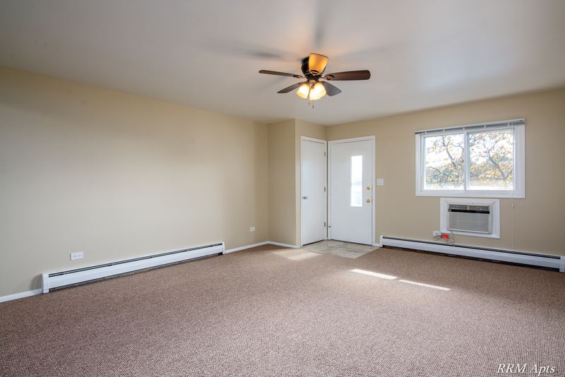 Living room with beige carpet, ceiling fan, entry door and a wall air conditioner under a window, Pistakee Road apartment 2B