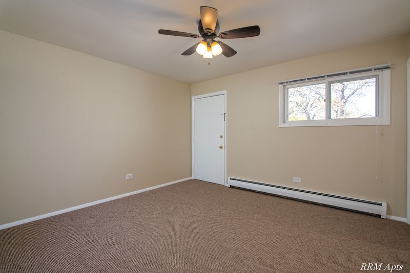 Empty room with beige carpet, ceiling fan, white door, window with trees and baseboard heater, Pistakee Road apartment 2B
