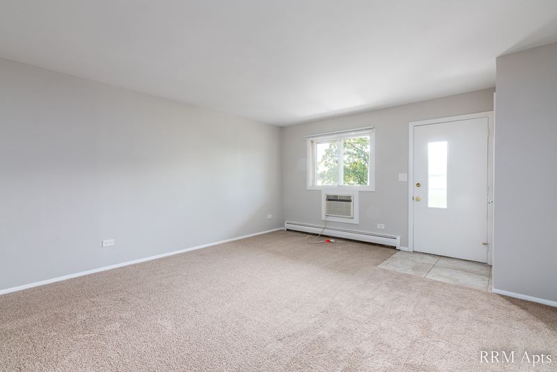 Living room with beige carpet, window over a wall air conditioner and an entry door, Pistakee Road apartment 2C