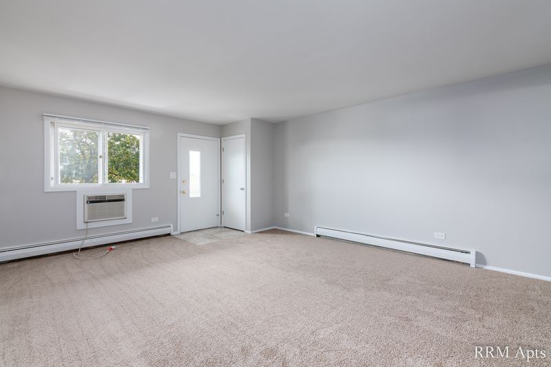 Living room with beige carpet, wall air conditioner under the window, entry door and hall, Pistakee Road apartment 2C