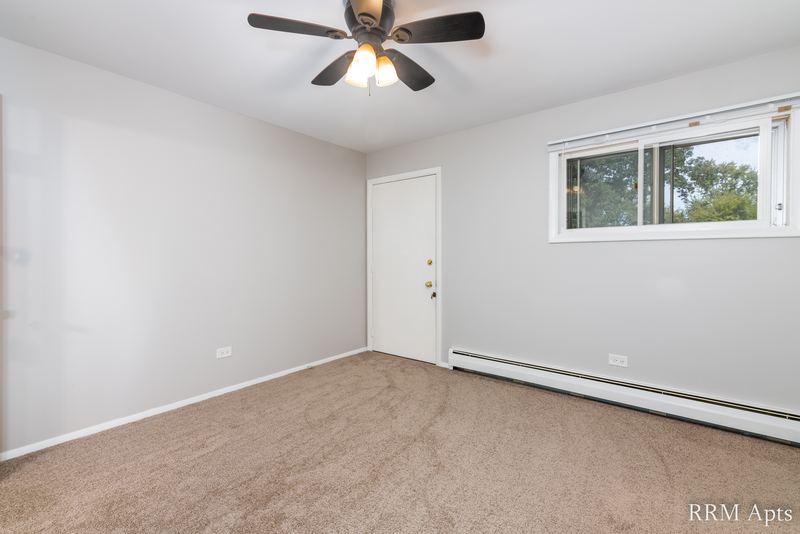 Empty room with beige carpet, ceiling fan, white door and a window onto trees, Pistakee Road apartment 2C