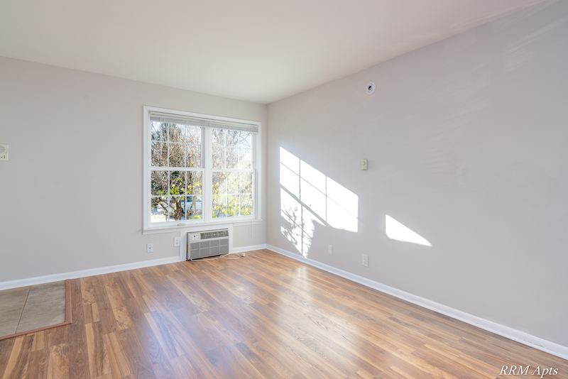 Living room with wood floor, sunlit window and a wall air conditioner below it, Prairie Ridge apartment 9716 1B
