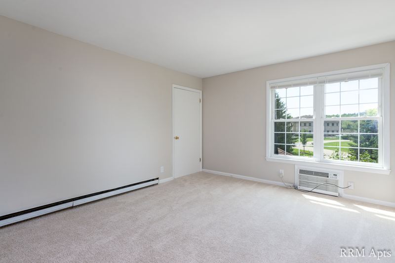 Living room with beige carpet, window over a wall air conditioner and baseboard heater, Prairie Ridge apartment 9716 2B