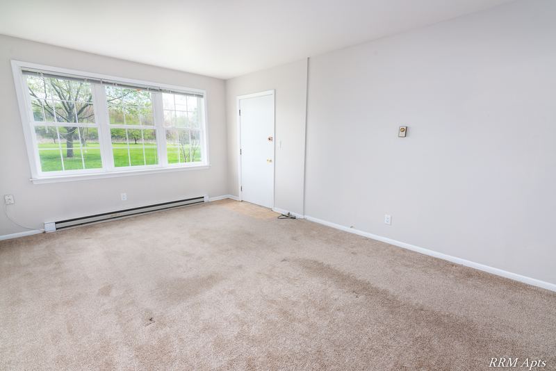Living room with beige carpet, wide window onto a lawn, baseboard heater and door, Prairie Ridge apartment 9716 GA