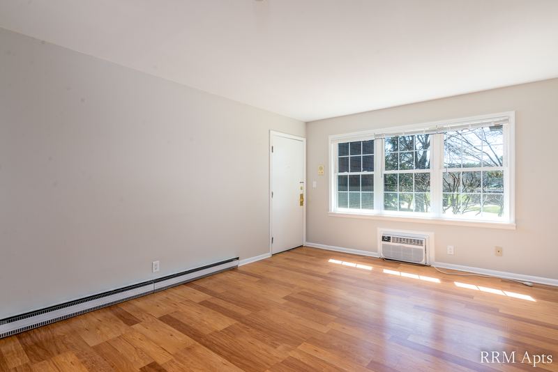 Living room with wood floor, window over a wall air conditioner and an entry door, Prairie Ridge apartment 9718-1D
