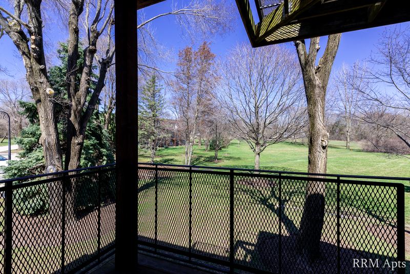 Balcony with black railing and a view of bare trees and lawn under a blue sky, Prairie Ridge apartment 9718-1D