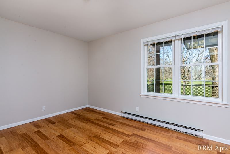 Empty room with wood floor, double window onto trees and a baseboard heater, Prairie Ridge apartment 9718-1D