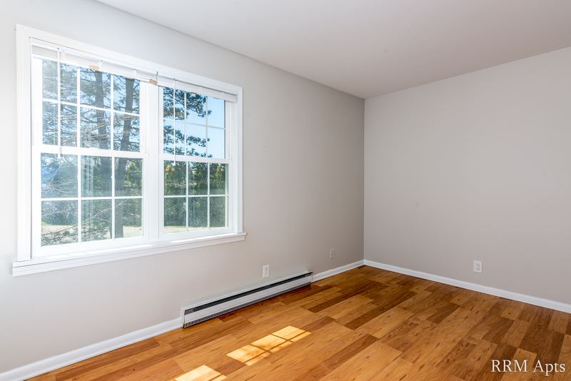 Empty room with wood floor, window onto trees on the left and a baseboard heater, Prairie Ridge apartment 9718-1D