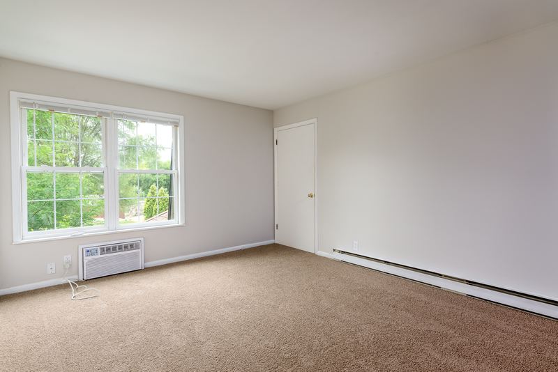 Living room with beige carpet, window over a wall air conditioner, baseboard heater and door, Prairie Ridge apartment 9718 2C