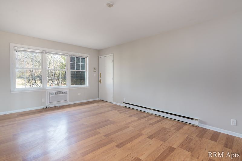 Living room with wood floor, wall air conditioner under a window, entry door and heater, Prairie Ridge apartment 9806 1A