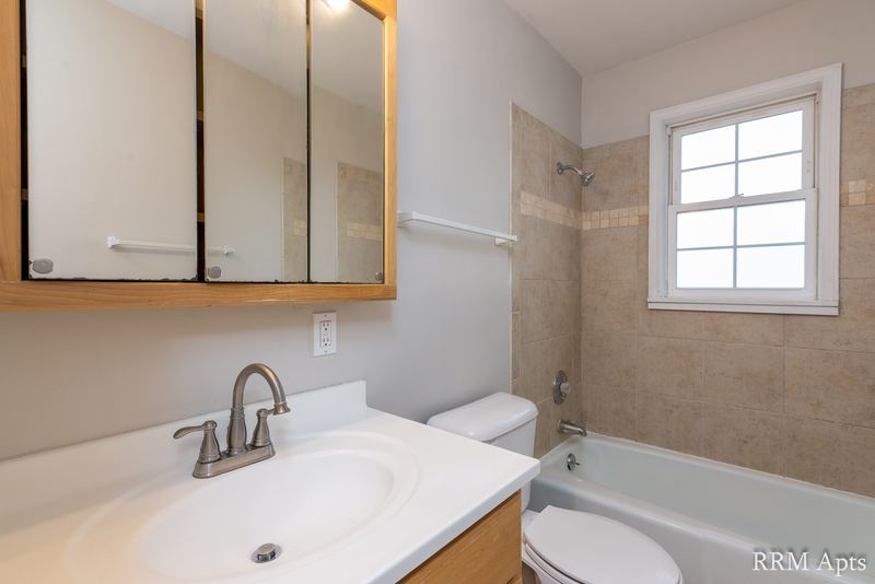 Bathroom with mirrored cabinet, white vanity, toilet and tub with tan tile and window, Prairie Ridge apartment 9806 1A