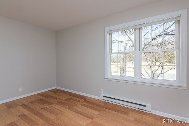 Empty room with wood floor, window onto bare trees on the right and baseboard heater, Prairie Ridge apartment 9806 1A