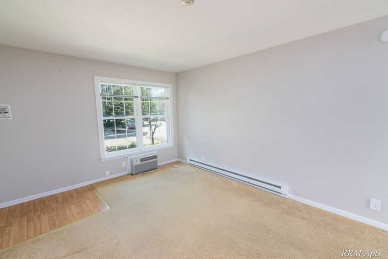 Living room with beige carpet, window over a wall air conditioner and baseboard heater, Prairie Ridge apartment 9806-1B