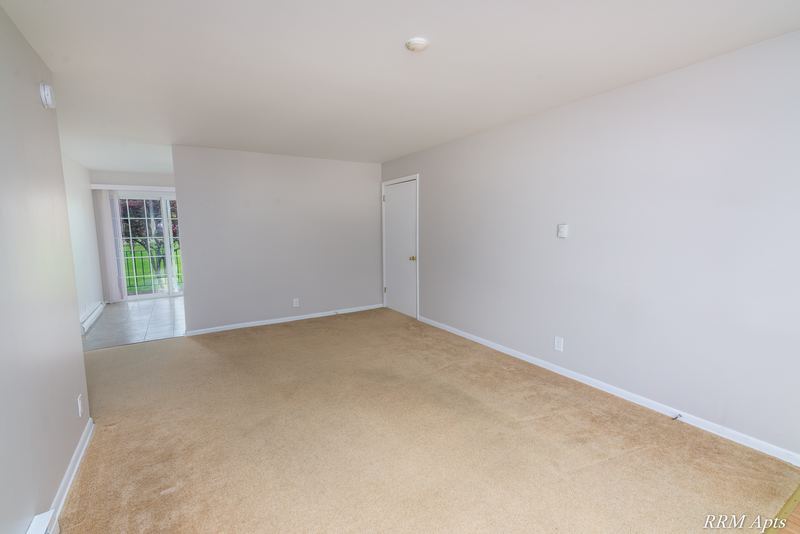 Living room with beige carpet, window on the left and an opening to the hall, Prairie Ridge apartment 9806-1B
