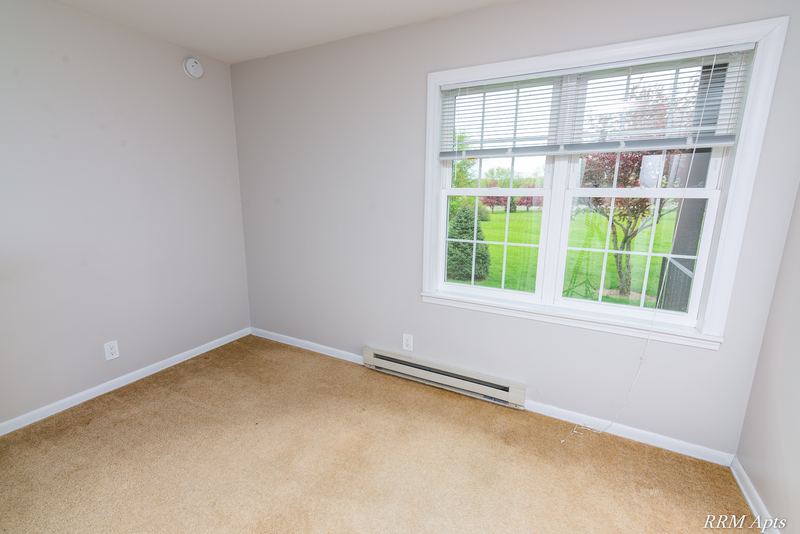 Empty room with beige carpet, wide window with blinds onto trees and baseboard heater, Prairie Ridge apartment 9806-1B