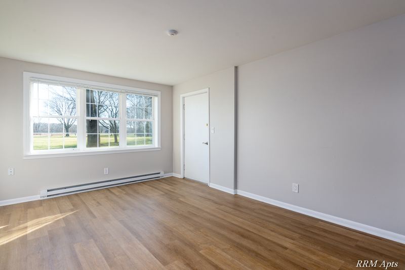 Living room with wood floor, window onto bare trees, entry door and baseboard heater, Prairie Ridge apartment 9806 GA