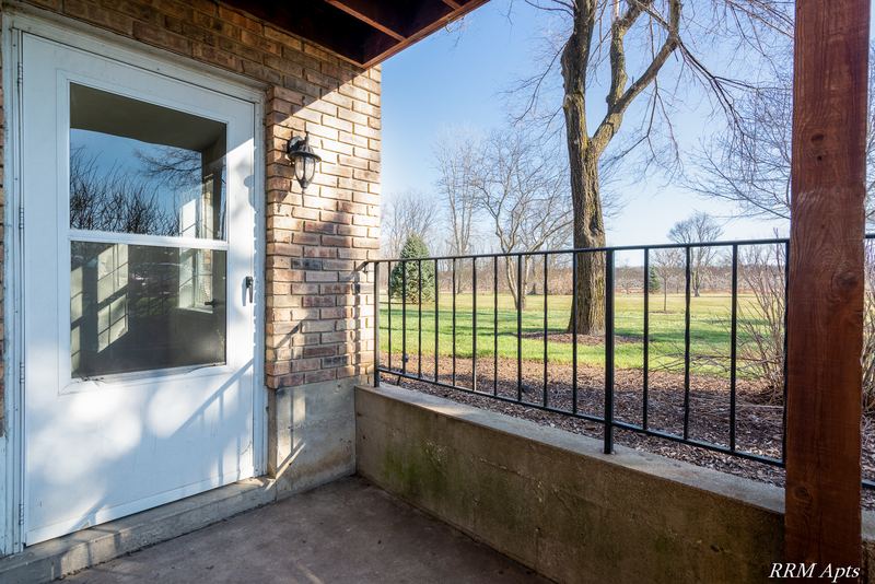 Patio with glass storm door, brick wall, black railing and a view of bare trees, Prairie Ridge apartment 9806 GA