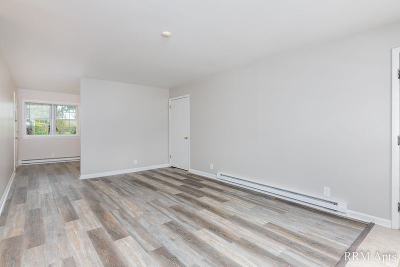 Living room with gray wood-look floor, window, baseboard heater and a hallway opening, Prairie Ridge apartment 9808-1D