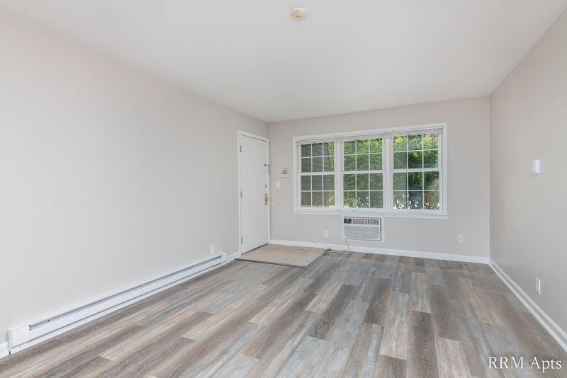 Living room with gray wood-look floor, window over a wall air conditioner and entry door, Prairie Ridge apartment 9808-1D