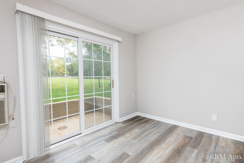 Living room with gray wood-look floor and a sliding glass door with curtain onto a lawn, Prairie Ridge apartment 9810 GB