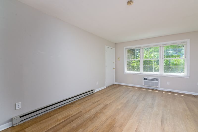 Living room with wood floor, wall air conditioner under the window and an entry door, Prairie Ridge apartment 9812 1D