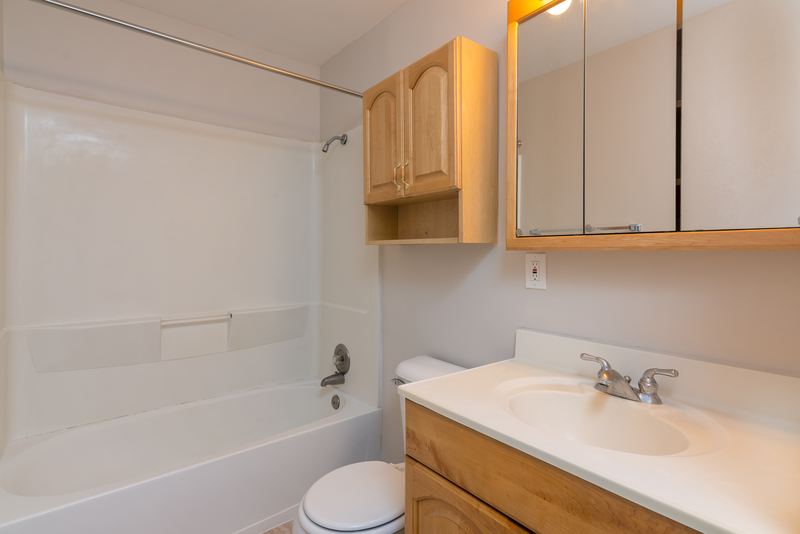Bathroom with white tub and surround, toilet, oak vanity and mirrored cabinet, Prairie Ridge apartment 9812 1D