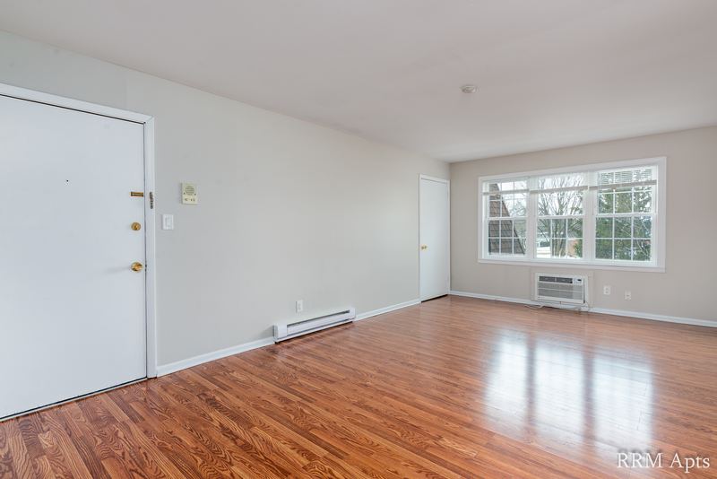 Living room with wood floor, entry door, window over a wall air conditioner and heater, Prairie Ridge apartment 9812 2D