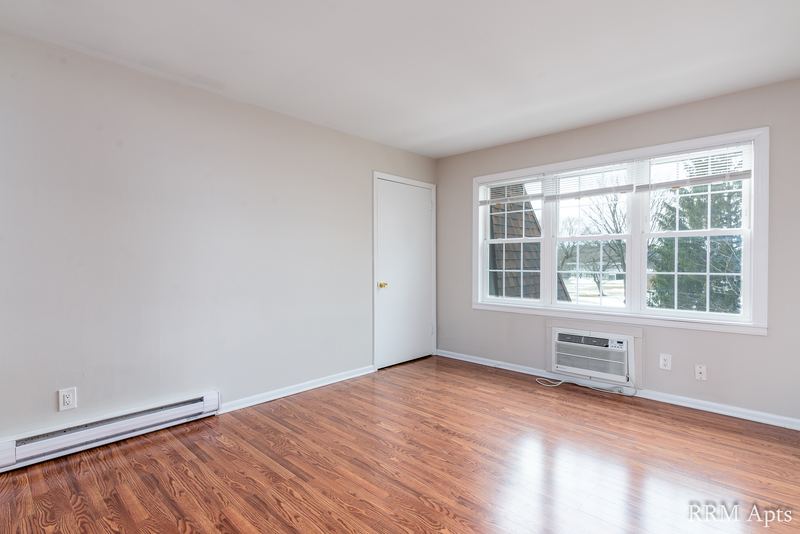 Living room with wood floor, white door and a wall air conditioner under the window, Prairie Ridge apartment 9812 2D
