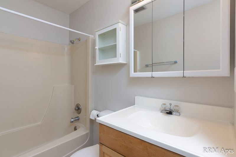Bathroom with white tub and surround, oak vanity, mirrored cabinet and small glass shelf, Prairie Ridge apartment 9812 2D