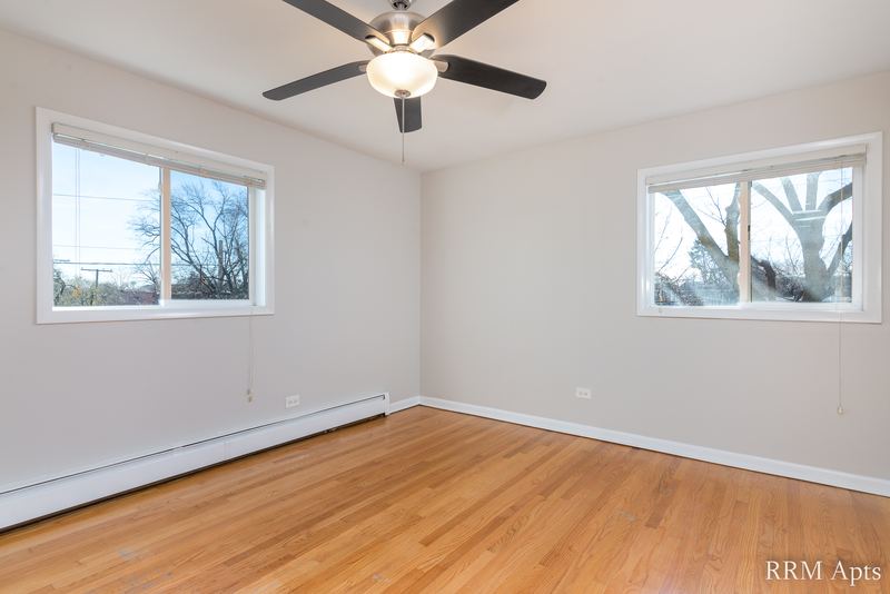 Empty room with wood floor, ceiling fan, two windows with trees and baseboard heater, Slusser Street apartment 6