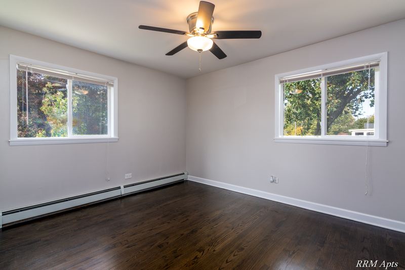 Bedroom with dark wood floor, ceiling fan, two windows facing trees and baseboard radiator, Slusser Street apartment 7