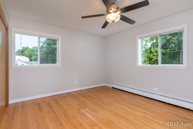 Bedroom with wood floor, ceiling fan, two windows facing trees and baseboard radiator, Slusser Street apartment 8