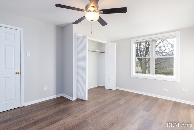 Bedroom with wood floor, ceiling fan, open closet, window facing trees and white door, South Slusser apartment 430