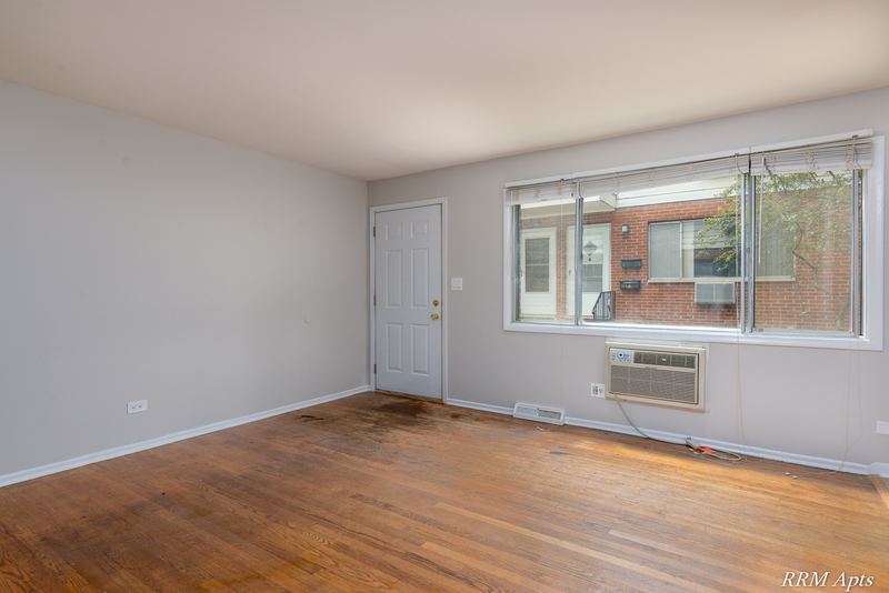 Living room with wood floor, window facing a brick building and wall air conditioner, Westerfield Place apartment 143A